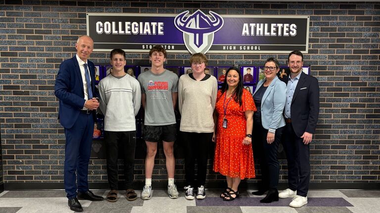 Eine Gruppe Personen steht vor einer Wand mit dem Banner "Collegiate Athletes Stoughton High School".