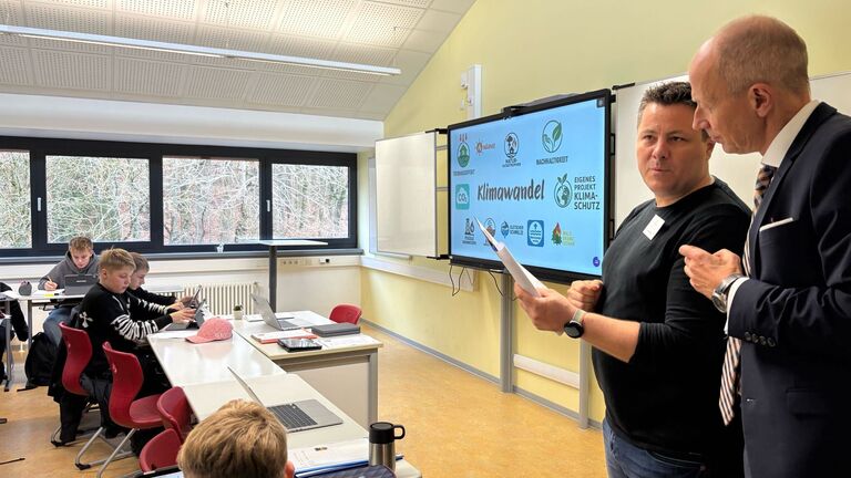 Schülerinnen und Schüler sitzen in einem Klassenraum und arbeiten mit Tablets und Laptops. An der Tafel ist eine Präsentation zum Thema Klimawandel mit Symbolen zu Nachhaltigkeit, CO₂ und Klimaschutz zu sehen.