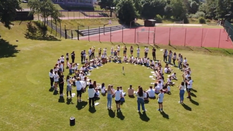 Schüler:innen stehen im Kreis auf einer Wiese (Luftaufnahme)