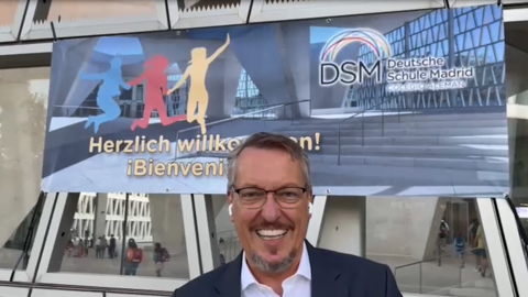 Schulleiter der Deutschen Schule Madrid, Dirk Pilgram, steht vor einem Banner und lächelt in die Kamera.