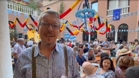 Schulleiter Dirk Pilgram steht in Tracht gekleidet vor einer Menge feiernder Menschen. Überall sind bunte Flaggen zu sehen.