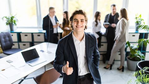 Junger Mann im Anzug steht in einem Büro und hält den Daumen hoch. Im Hintergrund unterhalten sich weitere Personen. Junger Mann im Anzug steht in einem Büro und hält den Daumen hoch. Im Hintergrund unterhalten sich weitere Personen.