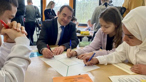Förderung für ukrainische Kinder und Jugendliche in Hessens Schulen Kultusminister Lorz sitzt gemeinsam mit Schülerinnen und Schülern um einen Tisch. Alle haben Stifte in der Hand und schreiben auf ein blatt Papier.