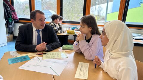 Förderung für ukrainische Kinder und Jugendliche in Hessens Schulen Kultusminister Lorz unterhält sich am Tisch sitzend mit zwei Schülerinnen, vor Ihnen liegen Aufgaben und Schreibmaterial. Im Hintergrund weitere Schüler an Gruppentischen.
