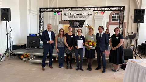 Staatssekretär Lösel auf der Veranstlatung vor einer Bühne stehend mit weiteren Personen.