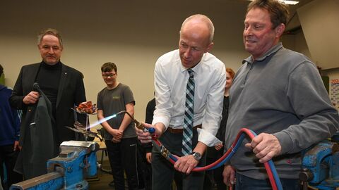 Bildungsminister Schwarz gemeinsam mit einem Herren an einem Brenner. Die Flamme auf eine Metallblume in einem Schraubstock gerichtet. 