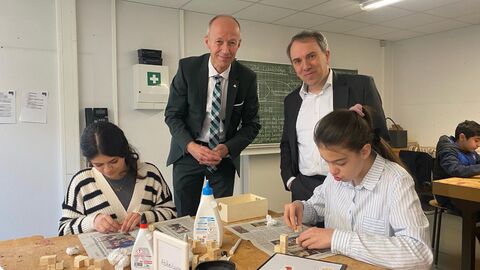 Bildungsminister Schwarz und der Vorsitzende der Geschäftsführung der Regionaldirektion Hessen der Bundesagentur für Arbeit, Dr. Frank Martin stehen hinter einem Tisch und beobachten die Arbeit zweier Schülerinnen die an diesem Tisch Holzklötze leimen.