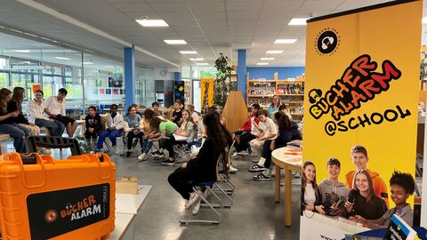 Bücheralarm @ school Blick in eine Schulklasse, die im Kreis sitzt. Im Vordergrund ein Aufsteller von "BÜCHERALARM @ school" sowie ein oranger Koffer mit der Aufschrift "Bücheralarm".