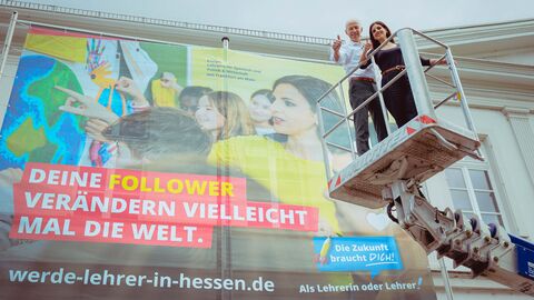 Initiative „Die Zukunft braucht Dich! Als Lehrerin oder Lehrer“ Von rechts auf einem Kran steht Kultusminister Schwarz gemeinsam mit einer Dame, hintendran an der Wand ist ein Plakat zu sehen mit der Aufschrift: "Deine Follower verändern vielleicht mal die Welt." der Initiative „Die Zukunft braucht Dich! Als Lehrerin oder Lehrer“.