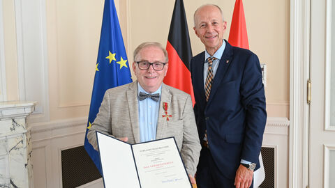 Staatsminister Schwarz steht gemeinsam mit Bertwin Spiller in einem Raum und überreicht eine Urkunde. Im Hintergrund stehen die Flaggen von Europa, der Bundesrepublik Deutschland und dem Land Hessen.