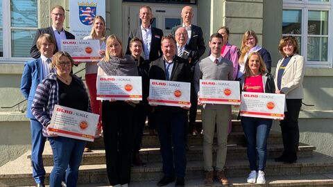 Eine Gruppe Personen steht vor einem Gebäude auf einer Treppe zum Gruppenfoto aufgestellt. Einige Personen halten große Checks in Händen.