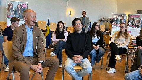 Armin Schwarz, Hessischer Minister für Kultus, Bildung und Chancen, im Gespräch mit Schülerinnen und Schülern der Friedrich-List-Schule Kassel über die EU. Der Minister sitzt auf einem Stuhl, umgeben von Schülern.