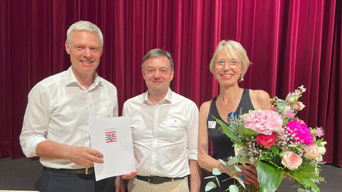 Staatssekretär Dr. Manuel Lösel gratuliert Dr. Matthias Bonczkowitz und der Projektleiterin Ruth Stücke aus dem Vorstand des Fördervereins Drei Personen stehen nebeneinander und halten einen Blumenstrauß und eine Urkunde.