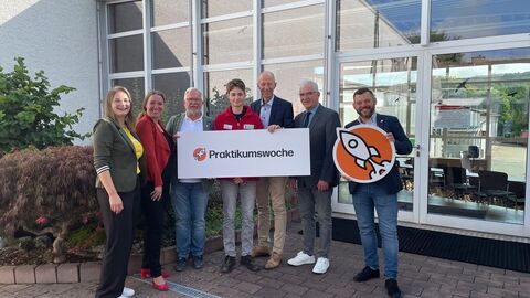 Mehrere Personen stehen vor Gebäude und halten Schild in Kamera.