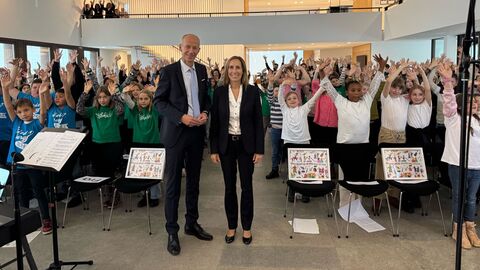 20 Jahre „Musikalische Grundschulen“ Zwei Erwachsene stehen in der Mitte. Im Hintergrund strecken viele Kinder die Hände in die Luft.