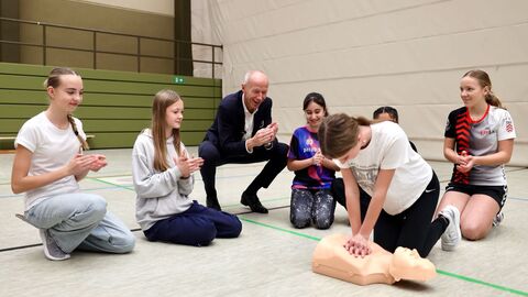 Eine Schülerin führt an einer Übungspuppe eine Herzdruckmassage durch. Weitere Schülerinnen sitzen im Halbkreis um sie herum, Bildungsminister Armin Schwarz unterstützt sie. Die Gruppe klatscht, um den Rhythmus vorzugeben.