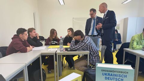 Der Botschafter und der Minister stehen in einem Klassenraum neben einem Klassentisch, an welchem 5 Schüler und Schülerinnen sitzen.