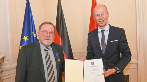 Zwei Männer stehen nebeneinander und halten eine Urkunde hoch. Im Hintergrund stehen eine Europa-Flagge, Deutschland-Flagge und Hessen-Flagge.