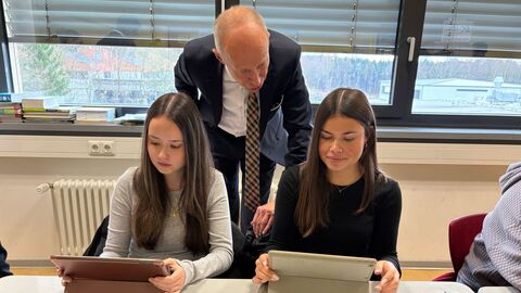 Zwei Schülerinnen sitzen an einem Tisch und schauen auf Tablets. Bildungsminister Schwarz steht hinter ihnen und schaut ihnen über die Schulter.