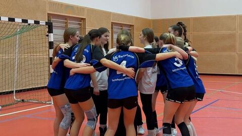 Eine Gruppe von Schülerinnen in Sportkleidung halten sich gegenseitig und stehen in einem Kreis in einer Sporthalle.