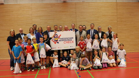 Bewegungscheck Grundschule Oberursel Sportminister Beuth und Kultusminister Lorz zwischen Kindern und Erwachsenen in einer Turnhalle. Sie stehen zum Gruppenfoto aufgestellt nebeneinander und halten ein Schild "Hessischer Bewegungs-Check" in den Händen.